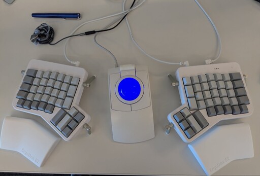 A white, split, ortholinear keyboard with no labels on the keys. Also, a white trackball mouse in the middle of the two keyboard halves with a blue ball in it.