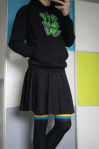 Photo of me standing in front of a wall, wearing a black skirt, a black hoodie with a green graffiti logo on it and black overknee socks with rainbow stripes.