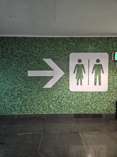 Wall with a bunch of green tiles in different hues. A sign leading to the bathroom